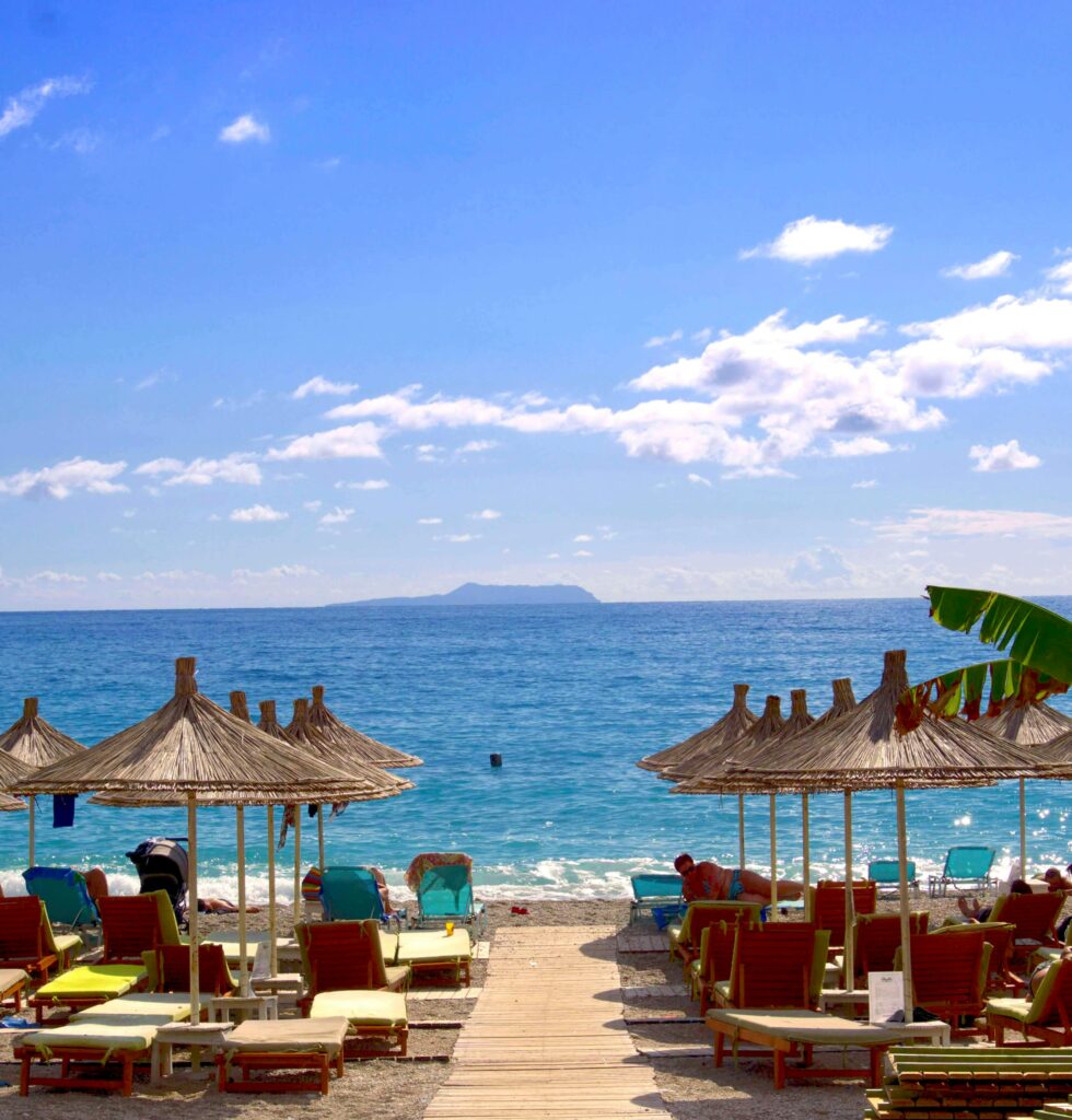 Home Relaxing summer day at Dhërmi beach, Albania with turquoise sea, umbrellas, and sun loungers.